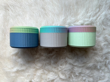 Three ribbed notions jars with pastel-colored lids and bodies, arranged side by side on a white fur background.