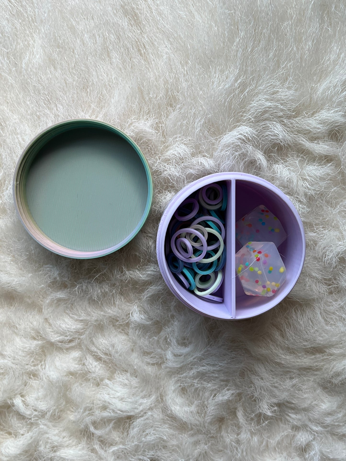 A pastel purple divided round organizer tray with a matching lid. The tray is open, showing colorful stitch markers and translucent stitch stoppers inside the compartments