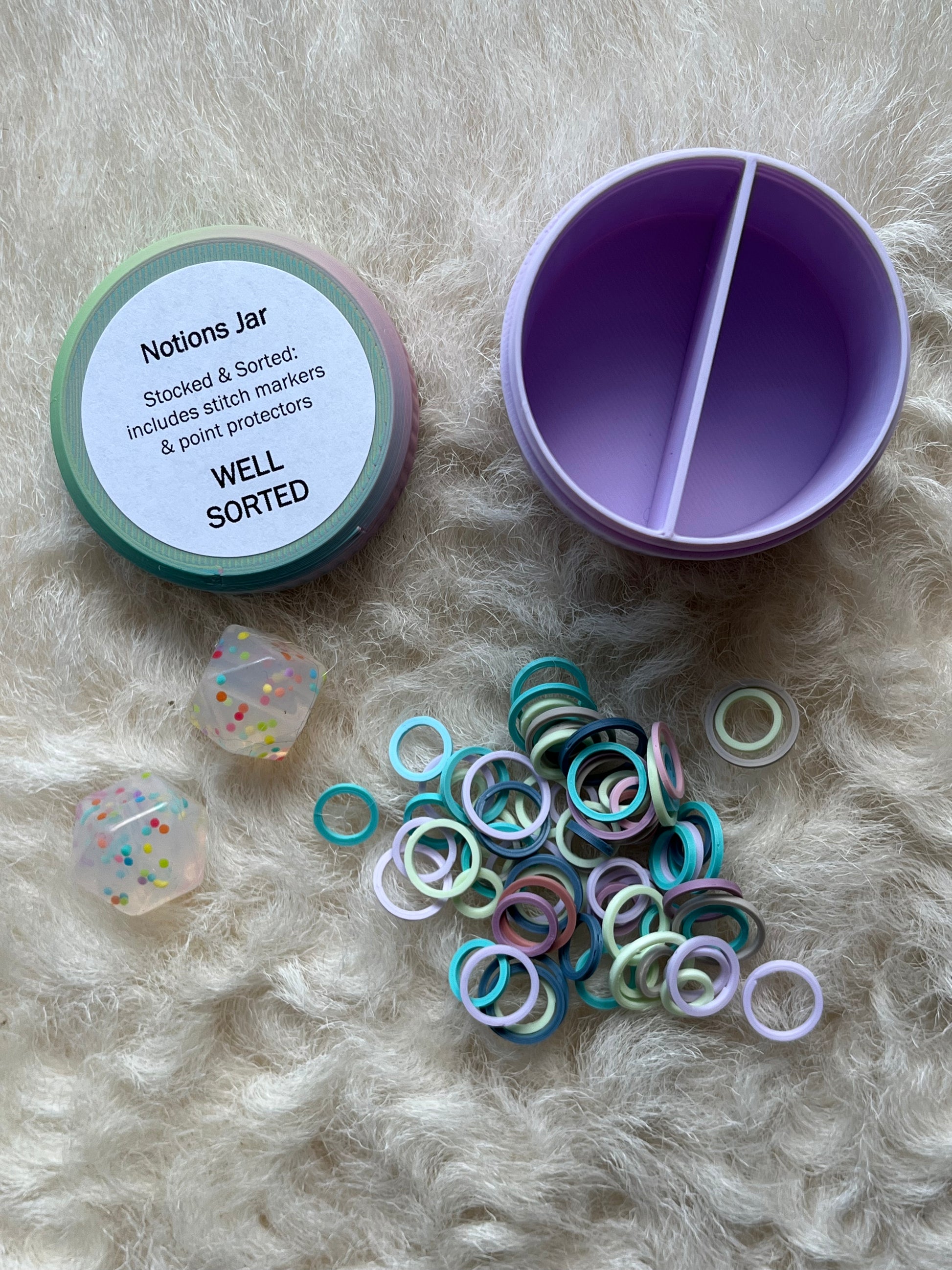 A pastel-colored 'Notions Jar' organizer shown open on a white fur background, alongside its lid, a pile of colorful stitch markers, and two clear stitch stoppers.