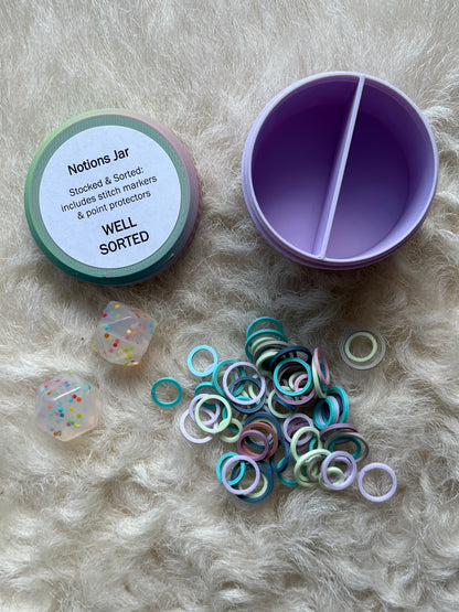 A pastel-colored 'Notions Jar' organizer shown open on a white fur background, alongside its lid, a pile of colorful stitch markers, and two clear stitch stoppers.