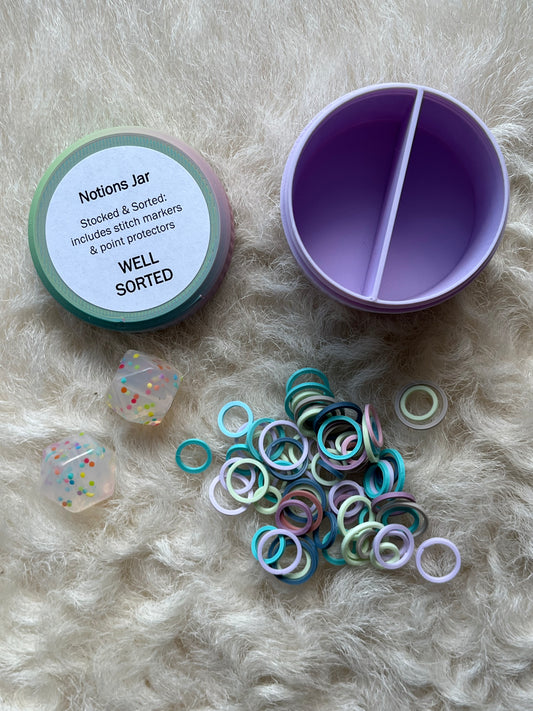 A pastel-colored 'Notions Jar' organizer shown open on a white fur background, alongside its lid, a pile of colorful stitch markers, and two clear stitch stoppers.