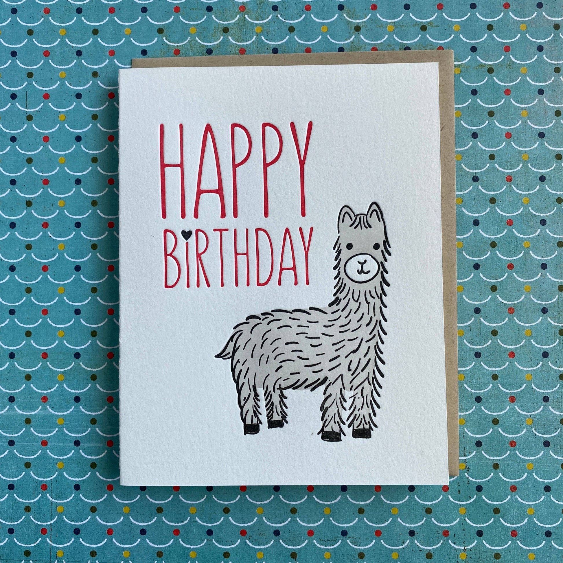 A white greeting card with a kraft envelope backing, featuring the words 'HAPPY BIRTHDAY' in red and a black-and-white alpaca illustration