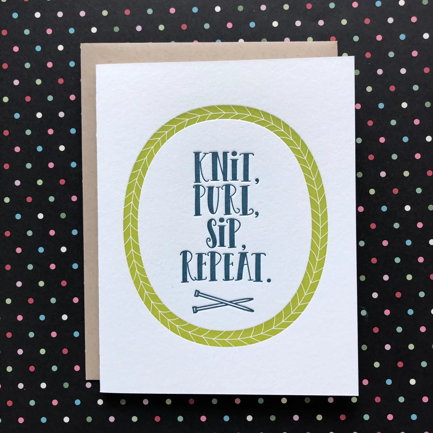 A white greeting card with a kraft envelope, featuring the text 'KNIT, PURL, SIP, REPEAT.' inside a light green oval border