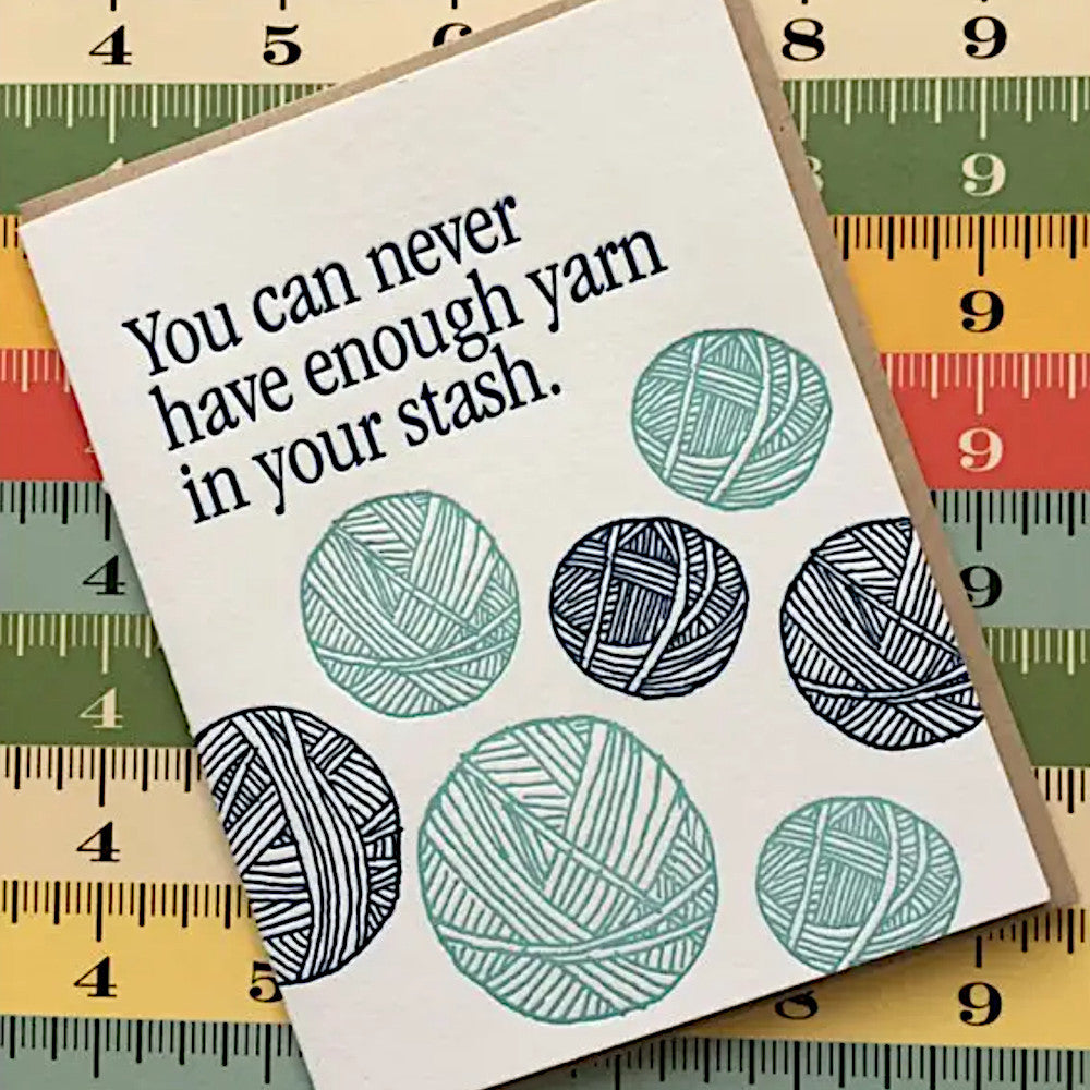 A square white card with the phrase 'You can never have enough yarn in your stash.' in black text and illustrated yarn balls in teal and dark navy