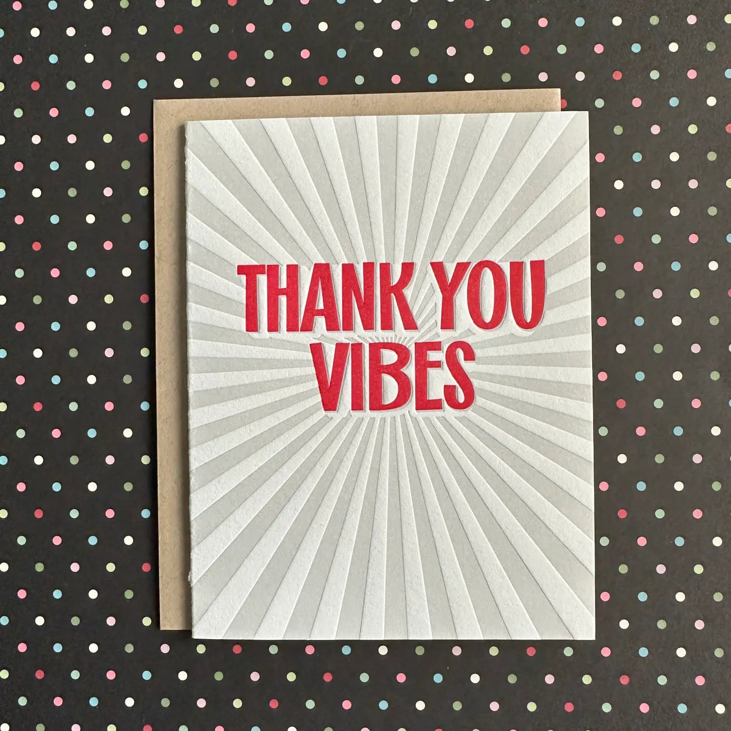 A square greeting card with a white sunburst design and the words 'THANK YOU VIBES' in bold red letters, placed on top of a brown envelope 