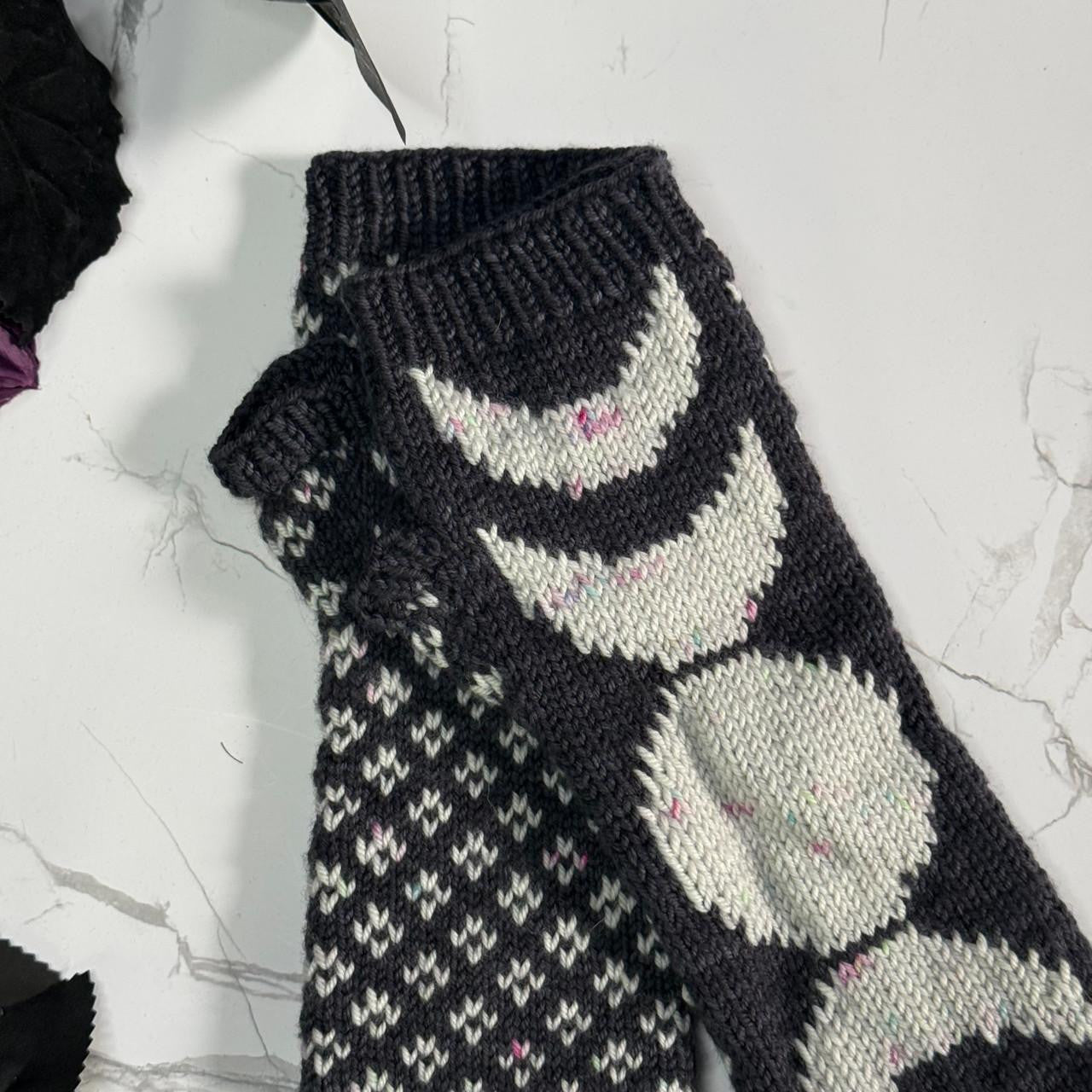 A close-up of a hand-knit dark gray and off-white fingerless mitten featuring the phases of the moon