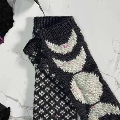 A close-up of a hand-knit dark gray and off-white fingerless mitten featuring the phases of the moon