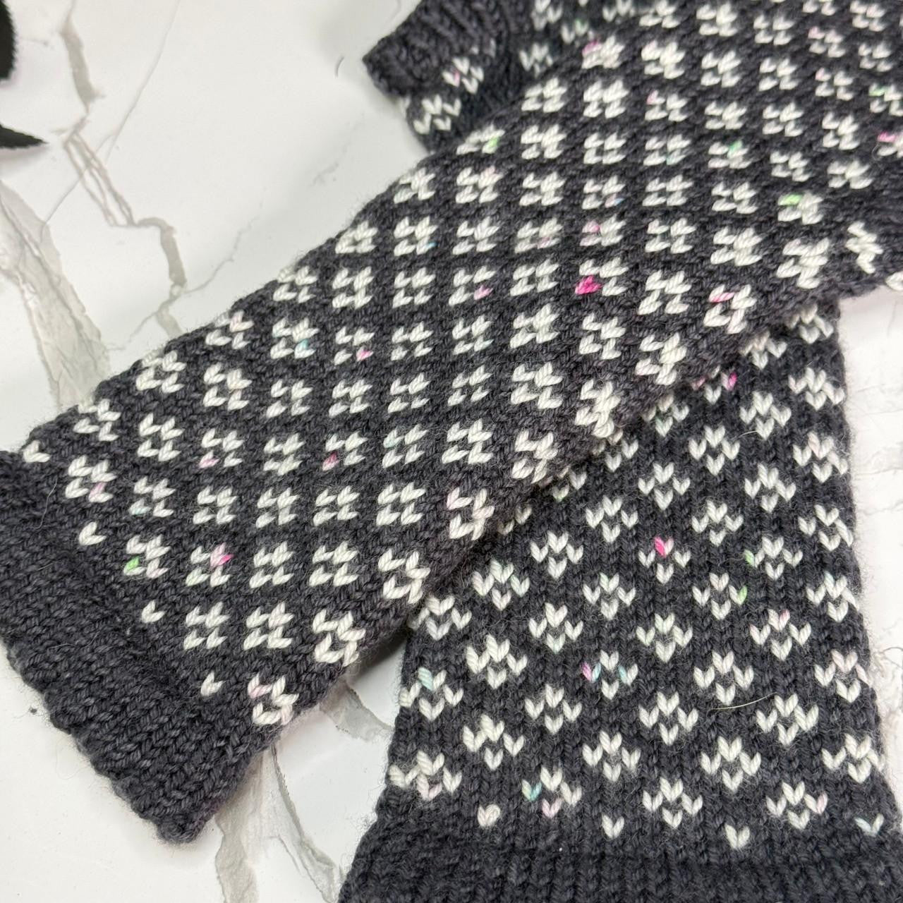 A close-up of a hand-knit dark gray and white patterned mitten laid flat on a light marble surface.