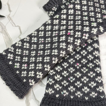 A close-up of a hand-knit dark gray and white patterned mitten laid flat on a light marble surface.